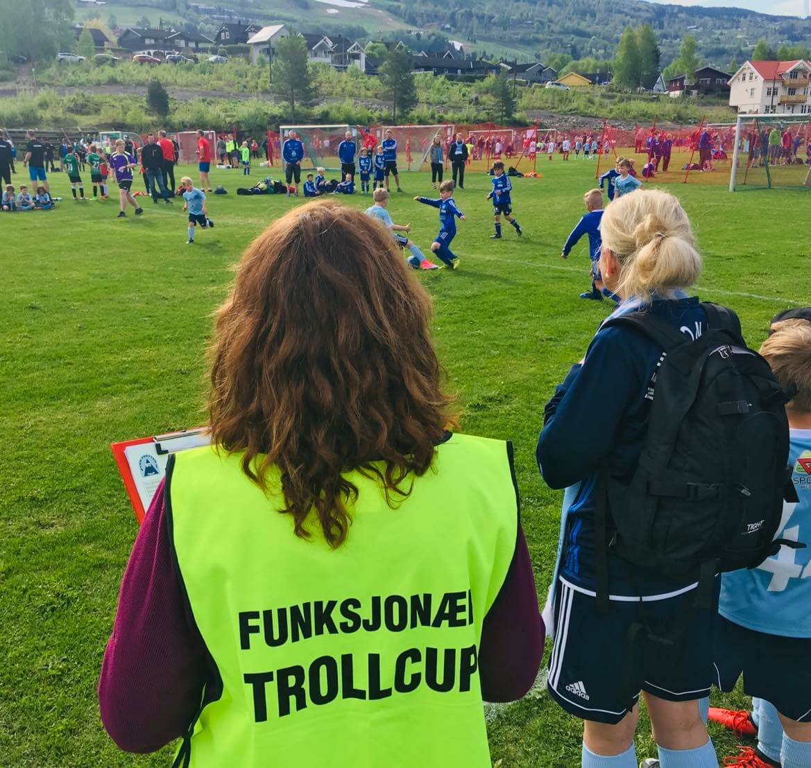 dame i gull vest med funksjonær på ryggen følger med på barnefotballkamp som dugnadsinnsats for klubb i Innlandet 